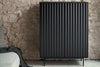 A tall black cabinet with vertical fluted doors and thin metal legs, shown in a styled interior setting against a textured stone wall.