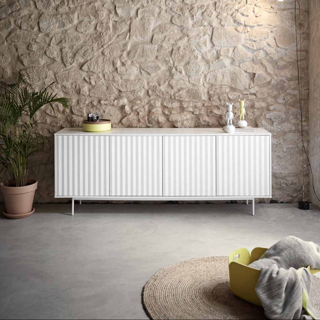 A modern white ribbed sideboard cabinet with slim legs, styled in a minimalist room with a textured stone wall, concrete floor, and a potted plant.