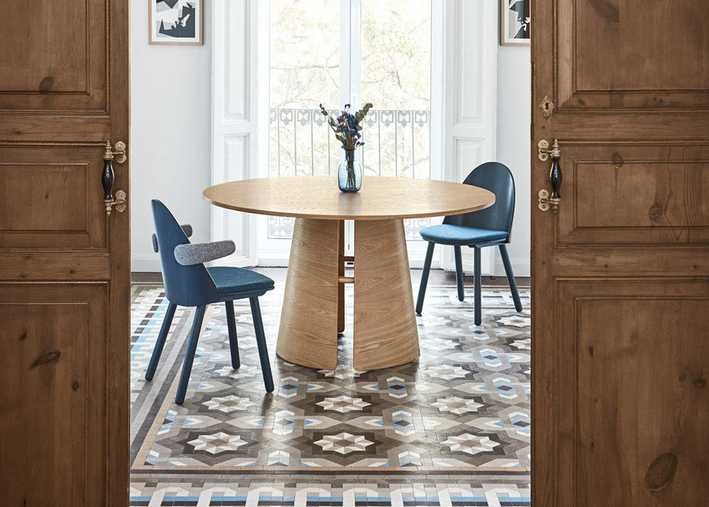 A round light wood dining table with a sculptural pedestal base, flanked by two blue chairs, set in a bright dining room viewed through open wooden doors.