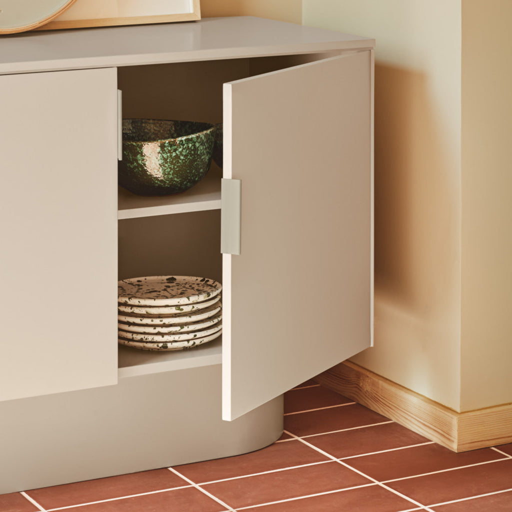 A modern light gray sideboard cabinet with one door open, styled in a warm beige room with terracotta tiled flooring. The cabinet is shown holding a green patterned bowl and stacked plates.