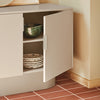 A modern light gray sideboard cabinet with one door open, styled in a warm beige room with terracotta tiled flooring. The cabinet is shown holding a green patterned bowl and stacked plates.