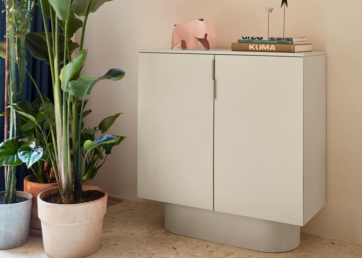 A sand finish two door cabinet from the Totem Occasional collection, placed in a room beside a plant pot.