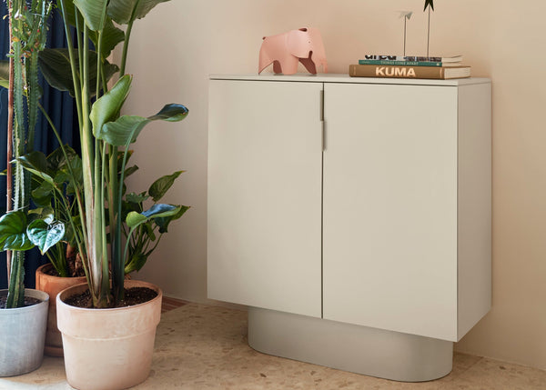 A sand finish two door cabinet from the Totem Occasional collection, placed in a room beside a plant pot.