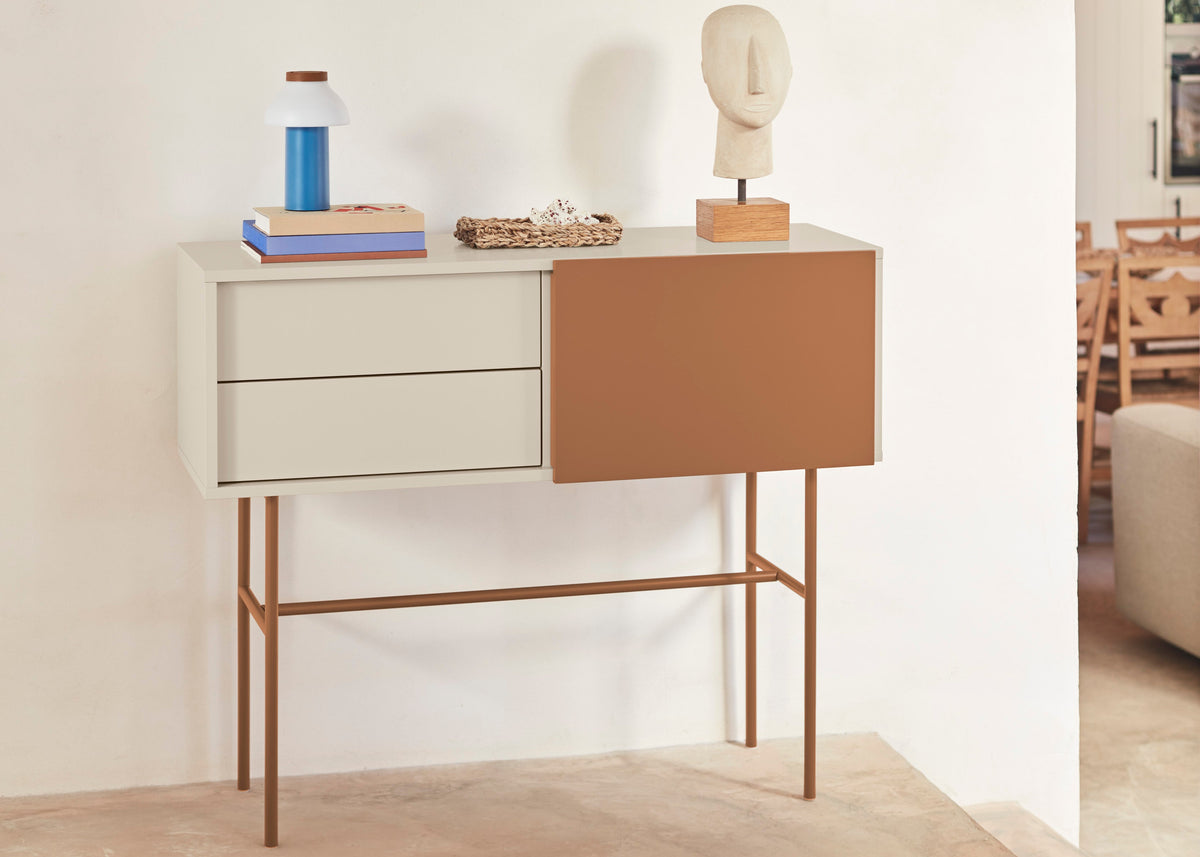 A mid-century modern console table with a white drawer section, a terracotta brown side panel, and bronze metal legs, styled with decorative objects in a minimalist room.