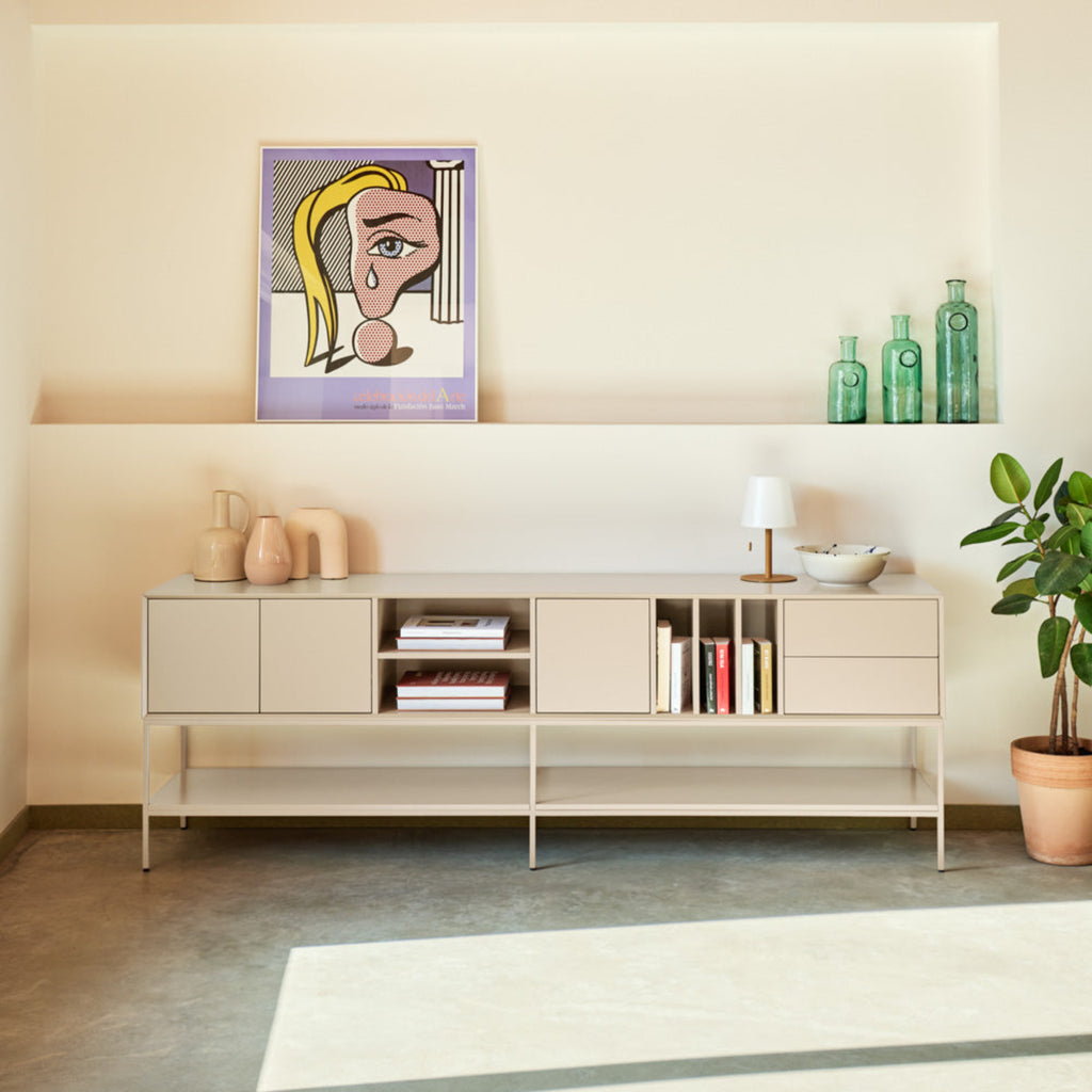A modern living room scene featuring a low-profile beige sideboard console with open shelving, styled with books, a small lamp, and decorative vases. Above the console, a framed pop-art portrait hangs on the cream wall.