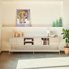 A modern living room scene featuring a low-profile beige sideboard console with open shelving, styled with books, a small lamp, and decorative vases. Above the console, a framed pop-art portrait hangs on the cream wall.