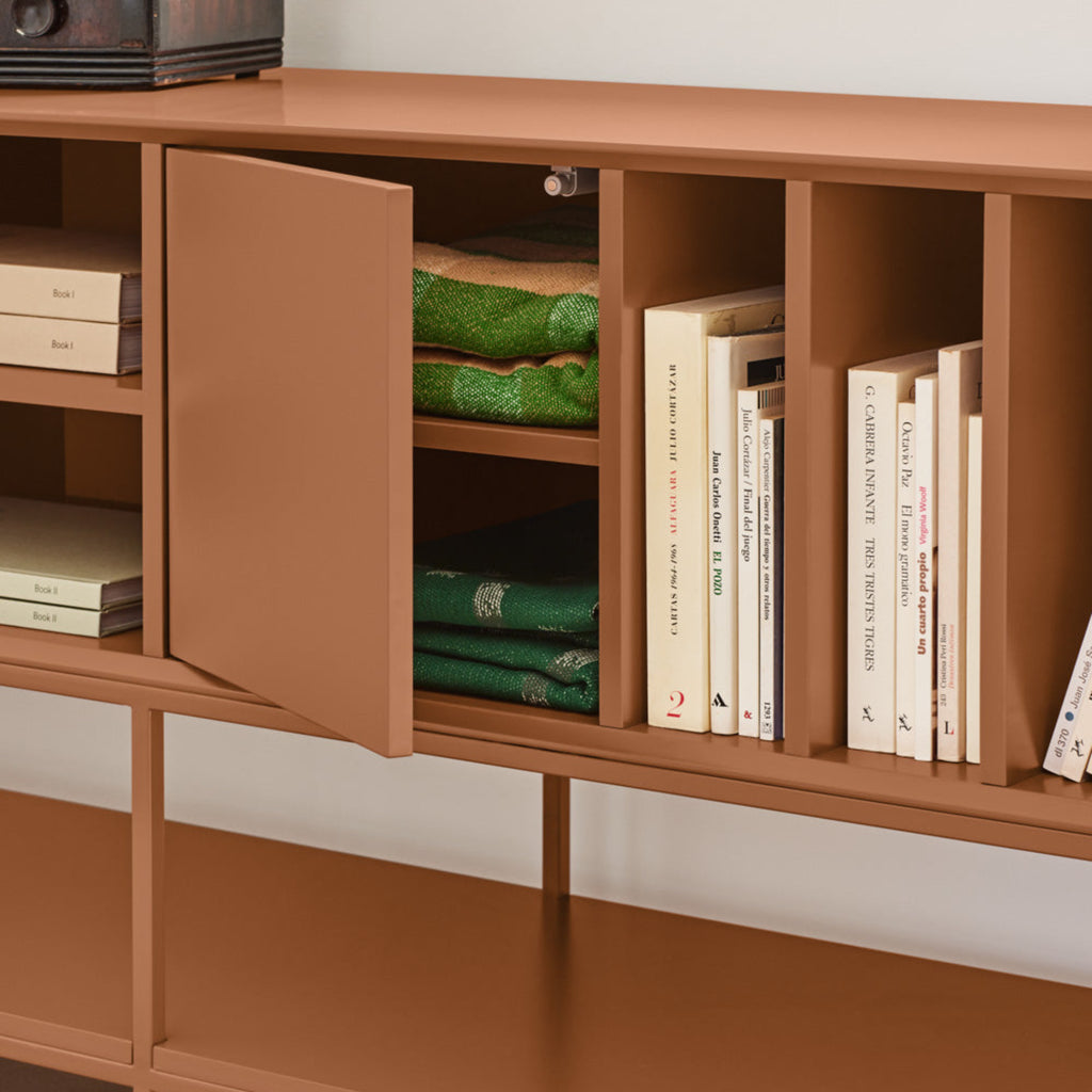 A mid-century modern wooden sideboard cabinet in a warm terracotta brown finish, shown with its door open to reveal folded green textiles and books inside. The cabinet is styled with books and green fabric, set against a neutral light gray wall and beige floor.