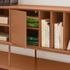 A mid-century modern wooden sideboard cabinet in a warm terracotta brown finish, shown with its door open to reveal folded green textiles and books inside. The cabinet is styled with books and green fabric, set against a neutral light gray wall and beige floor.