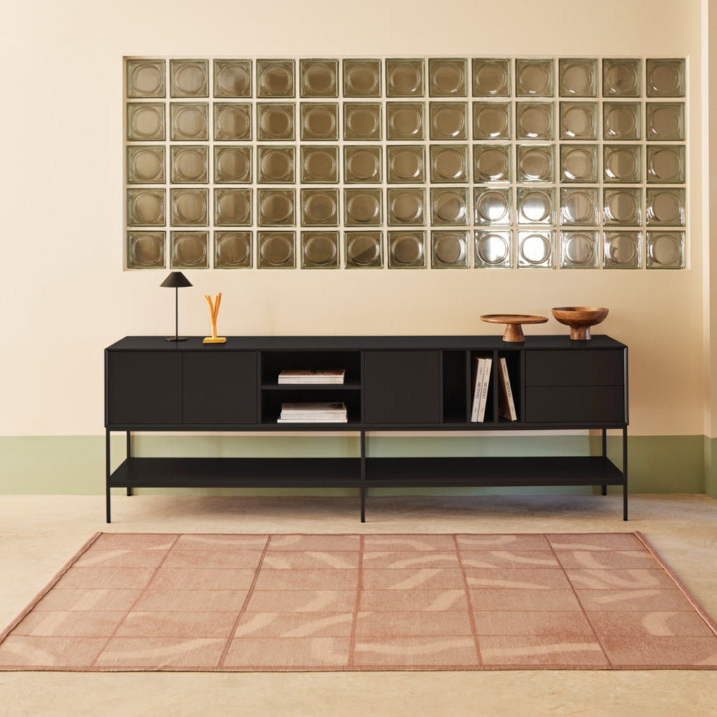 A modern black media console with open shelving and a lower shelf, styled in a minimalist room with a pink rug, a glass block wall, and a small table lamp.