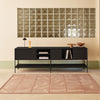 A modern black media console with open shelving and a lower shelf, styled in a minimalist room with a pink rug, a glass block wall, and a small table lamp.