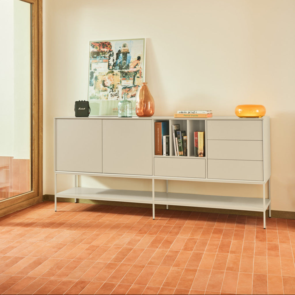 A modern white sideboard cabinet with open shelving and drawers, styled with books, a framed collage, and decorative vases in a minimalist room with terracotta tiled floors.