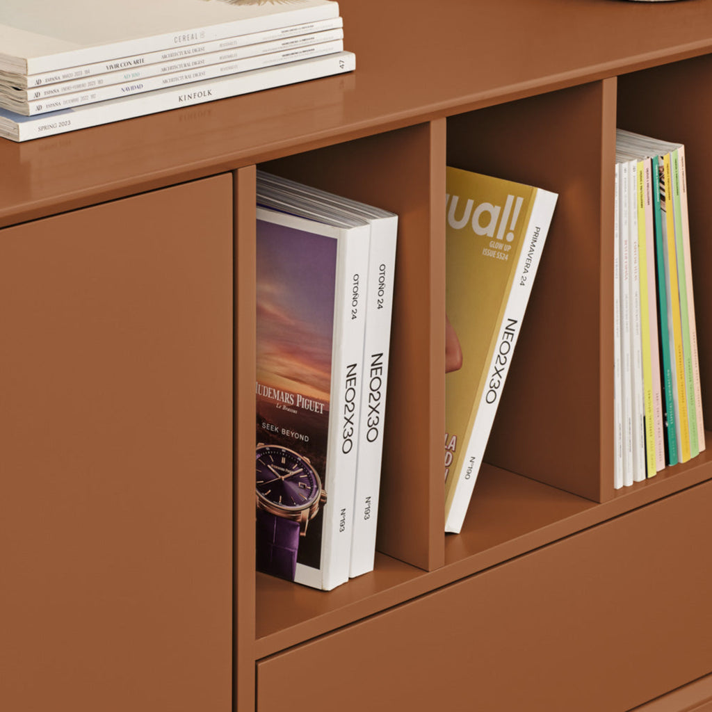 A close-up of a modern brown wooden media console with open cubbies and a drawer, styled with books and magazines, against a beige wall.