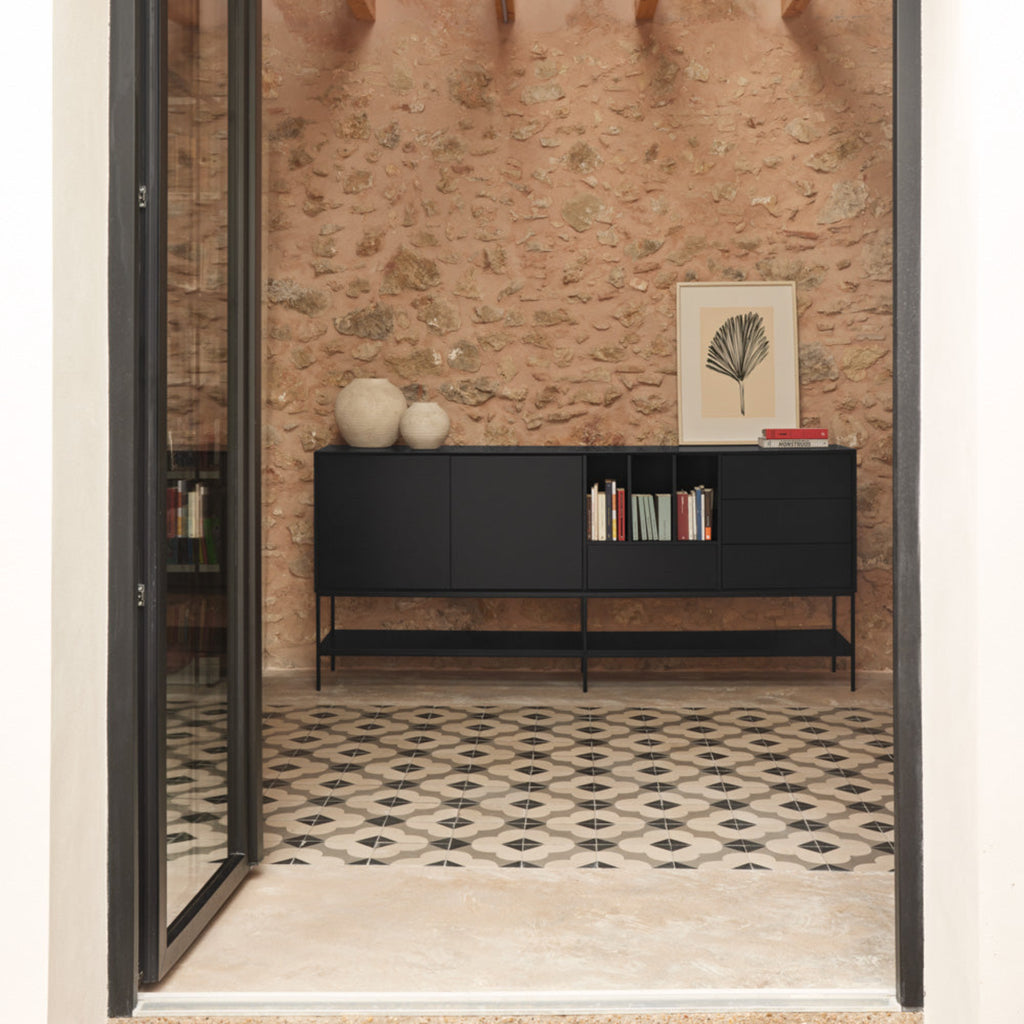 A modern black wooden sideboard cabinet with open shelving, styled in a rustic room with a stone wall, patterned tile floor, and exposed wooden ceiling beams.