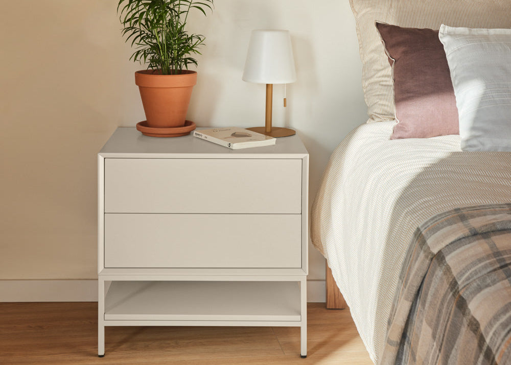 A modern white two-drawer nightstand with an open lower shelf, styled in a bedroom next to a bed with white linens and pillows, featuring a potted plant and a table lamp on top.