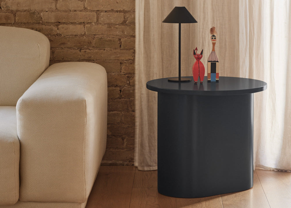 A modern round side table with a matte black finish, styled in a living room next to a cream-colored armchair, featuring a black table lamp and two small decorative figurines on top.