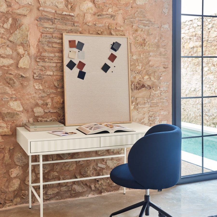 A white Sierra two-drawer desk with a minimalist design, placed against a brick wall backdrop with a blue chair in front of it.